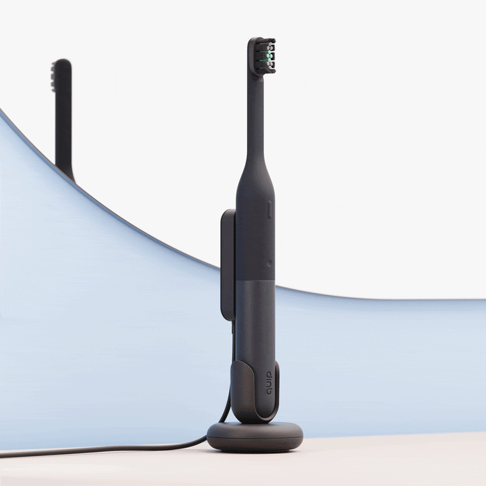 A sleek black electric toothbrush standing upright on a matching charging base, set against a soft blue and white curved background with its reflection visible behind it.