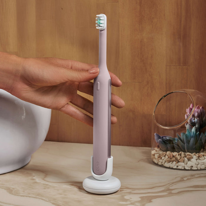 Hand placing a pink electric toothbrush onto its white charging stand on a bathroom counter.